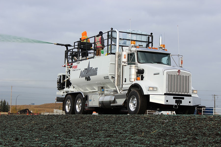 Two people on top of a white Finn Hydroseeder attached to a white truck as it sprays a field.