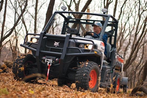 Man in blue hat driving a white Bobcat® UTV through a forest full of dead leaves on the ground.